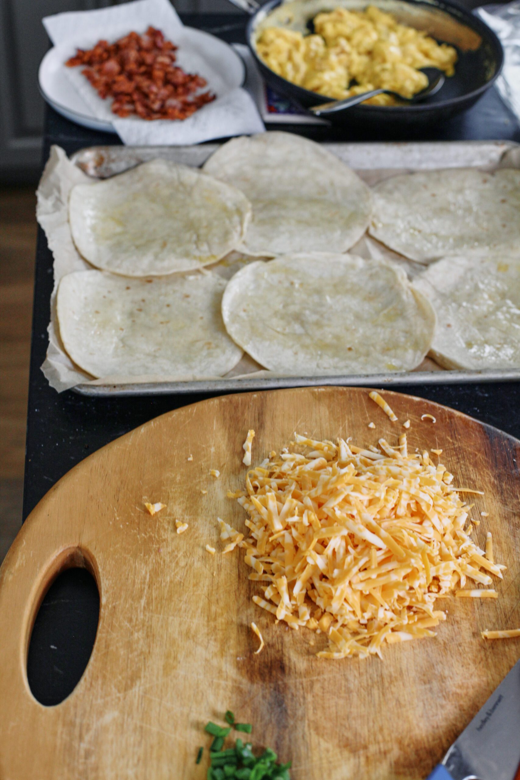 Bacon and Egg Sheet Pan Tacos 3 A frying pan with scrambled eggs, a plate of chopped, cooked bacon, tortillas on a sheet pan, and shredded cheese on a cutting board.