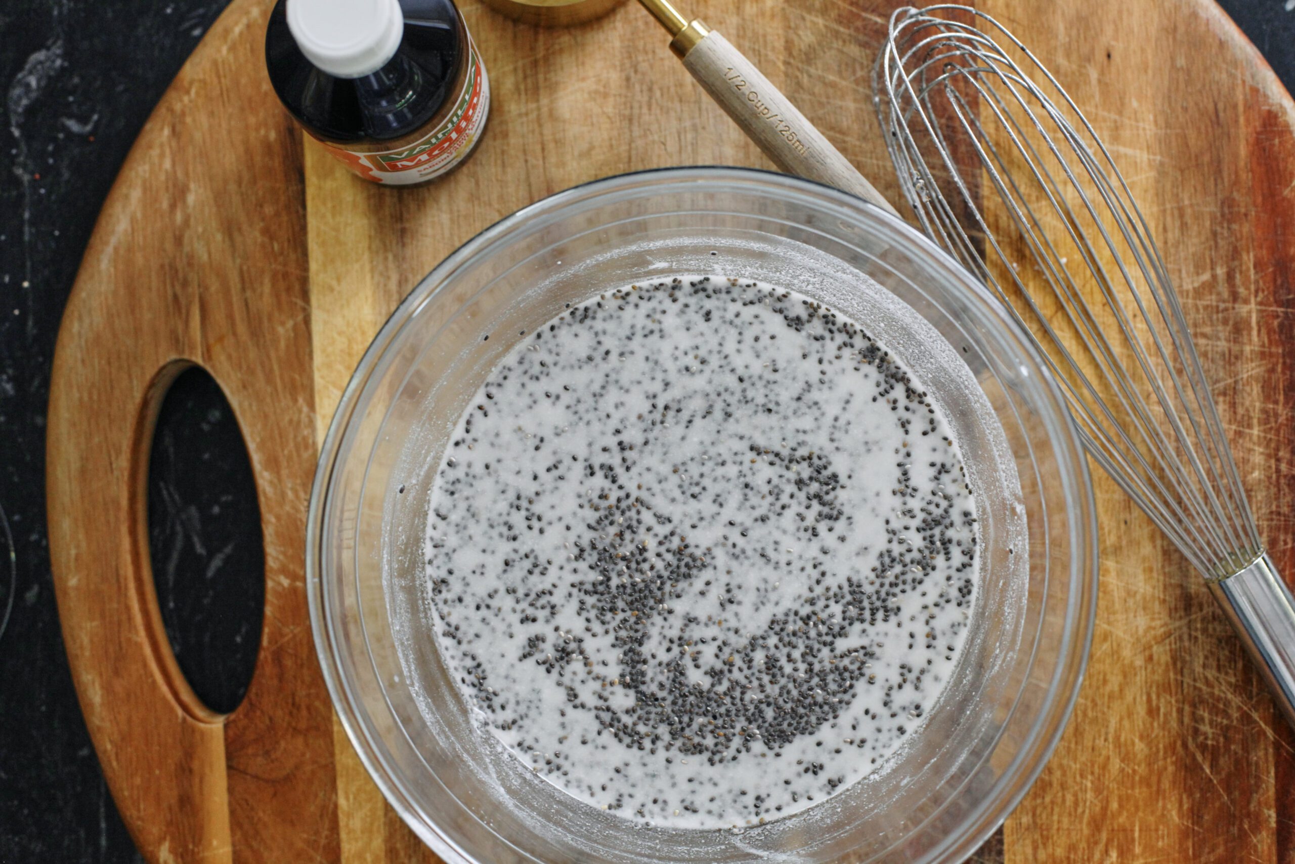 A glass bowl on a cutting board with coconut milk and chia seeds in it. There's also a bottle of vanilla extract, a measuring cup, and a whisk on the cutting board.