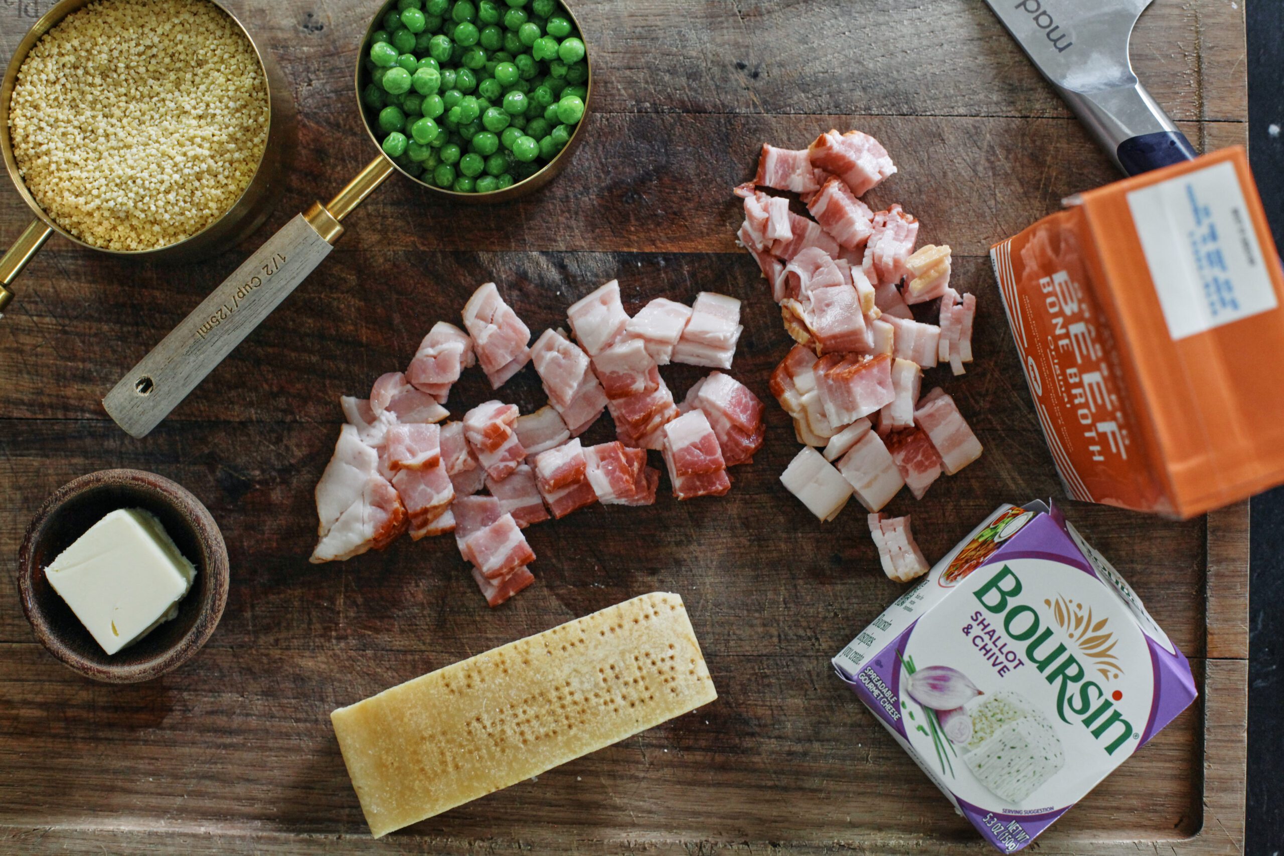 ingredients prepped on a wooden cutting board with a knife in the frame: pastina in a 1 cup measuring cup, peas in a 1/2 cup measuring cup, a knob of butter in a small bowl, chopped bacon, chunk of parmesan cheese, boursin shallot & chive cheese, carton of beef broth
