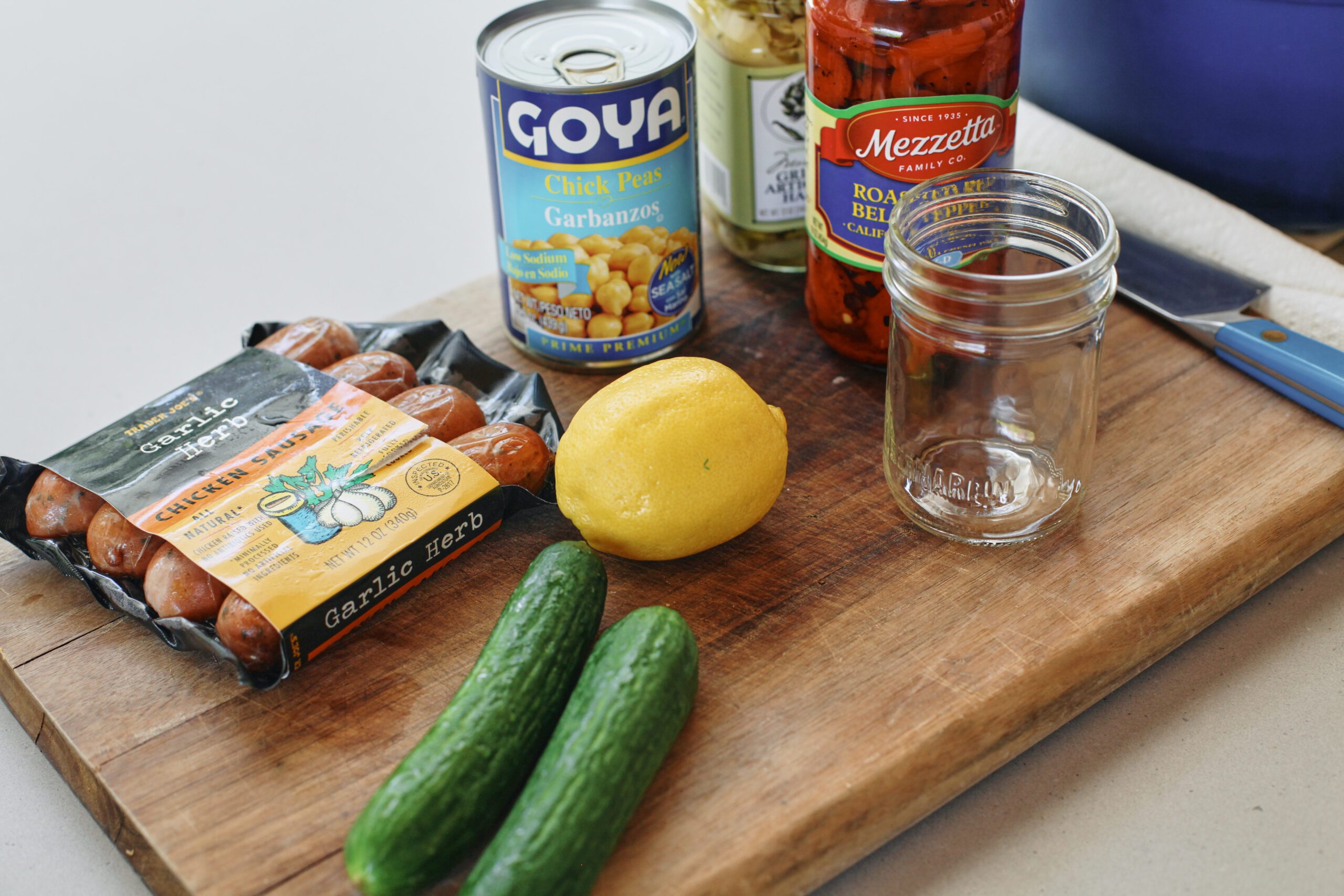 ingredients prepped for high protein pasta sald: sausages, cucumbers, lemon, can of chickpeas, jarred marinated artichokes, jarred roasted red peppers, and a glass jar for the dressing