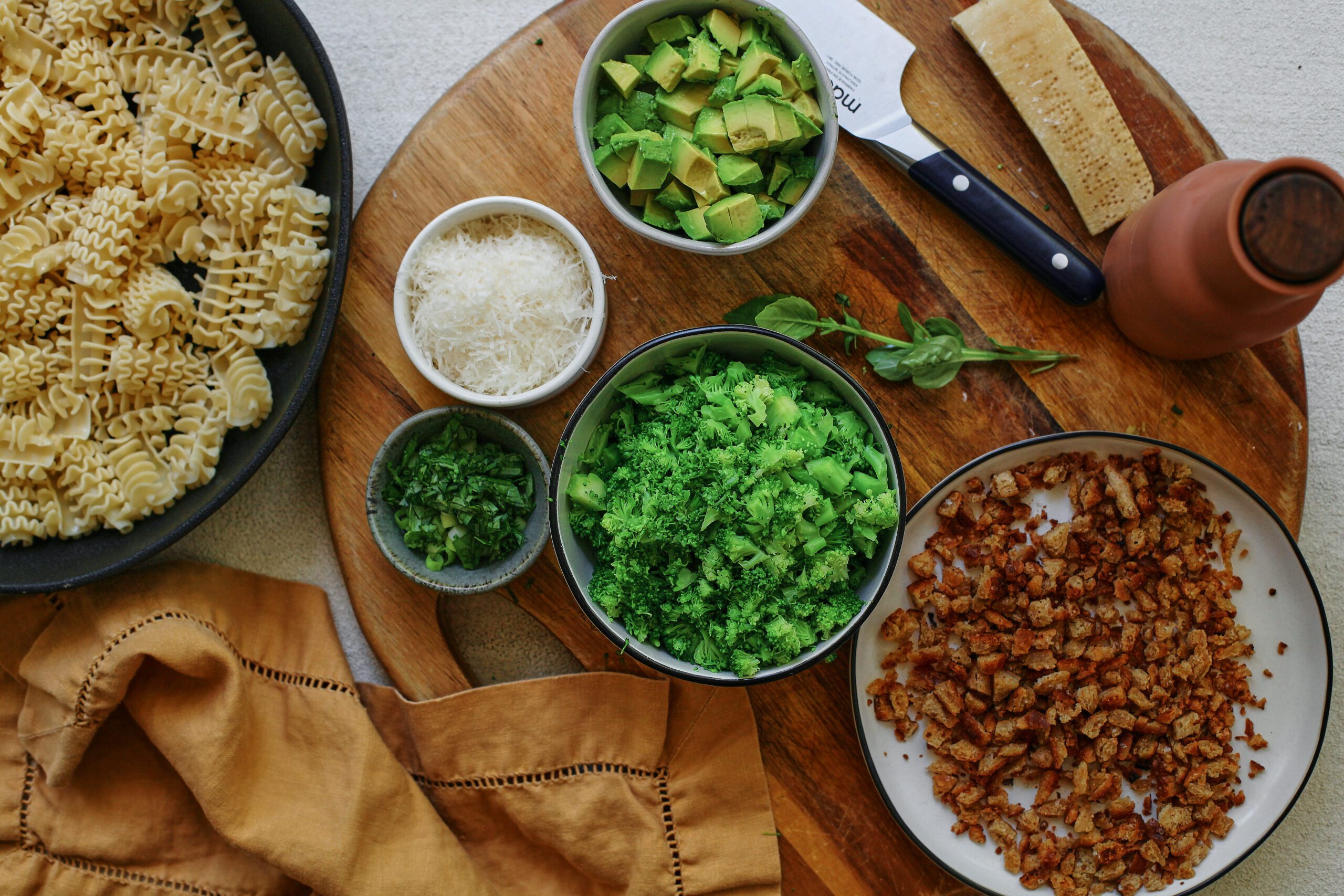 prepped ingredients for broccoli caesar pasta salad: cooked pasta, chopped broccoli, avocado, green onions, and basil, grated parmesan cheese, toasted bread crumbs