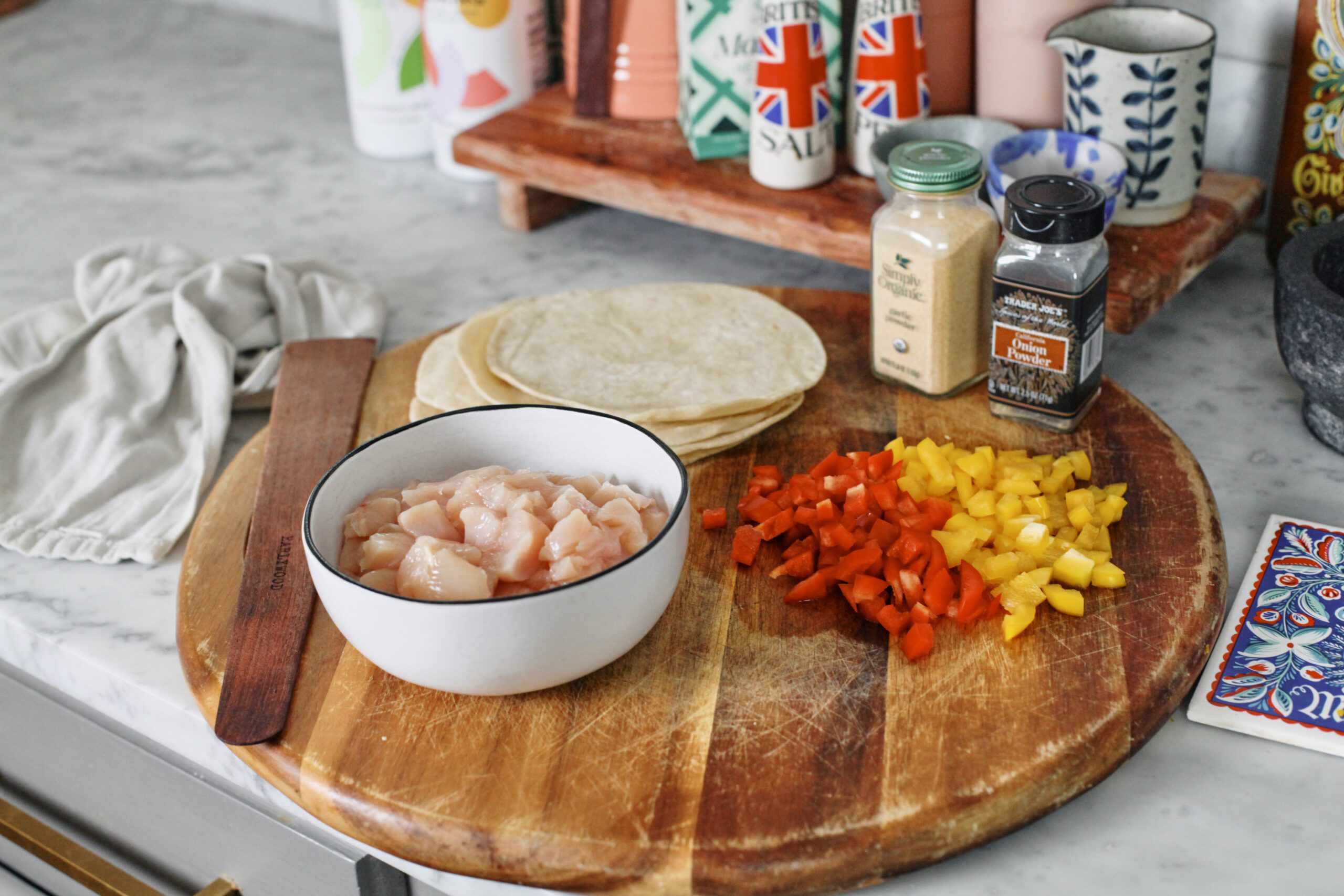 ingredients for buffalo chicken sheet pan tacos prepped on a wood cutting board: chopped raw chicken breasts in a bowl, tortillas, chopped yellow and red bell pepper, garlic powder, onion powder