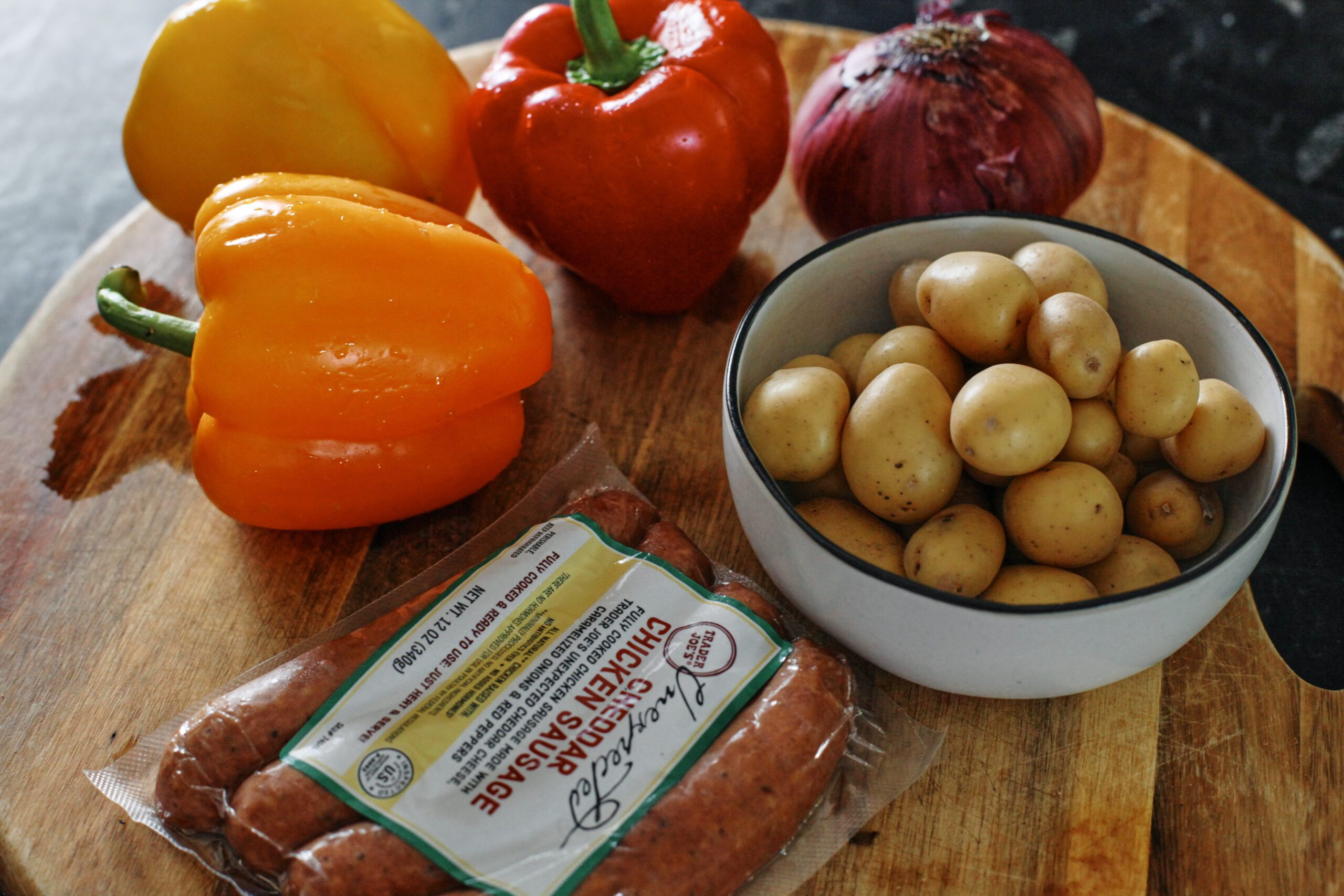 ingredients on a cutting board: bell peppers, red onion, baby poatoes, a package of chicken sausages