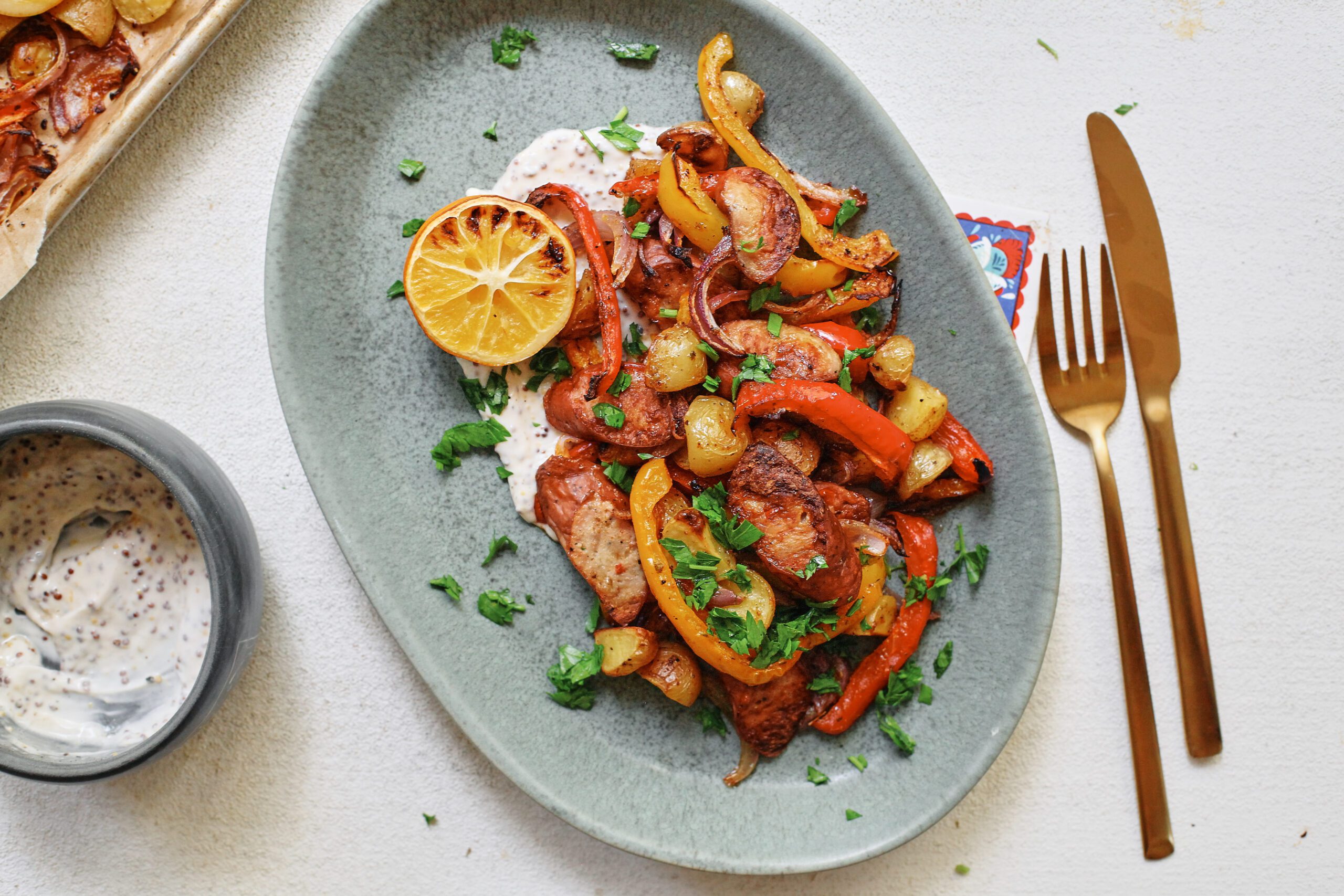 sheet pan sausage, peppers, and potatoes served on a blue plate over a white sauce with half a roasted lemon