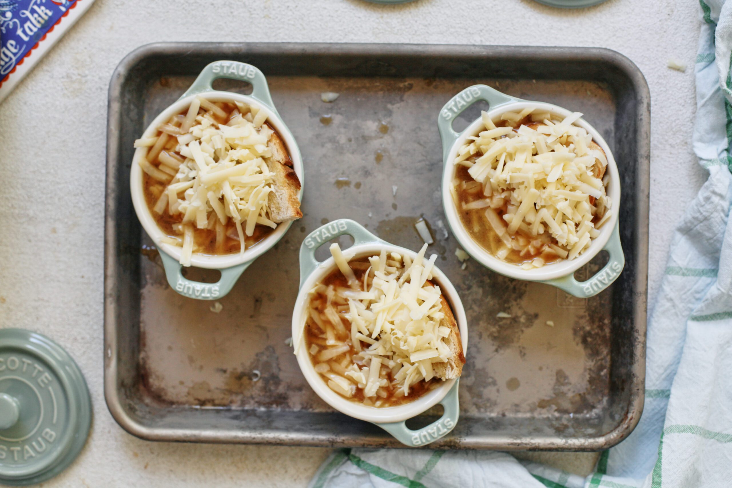 french onion soup in mini green dutch ovens that are arranged on a sheet pan, topped with shredded cheese before going under the broiler
