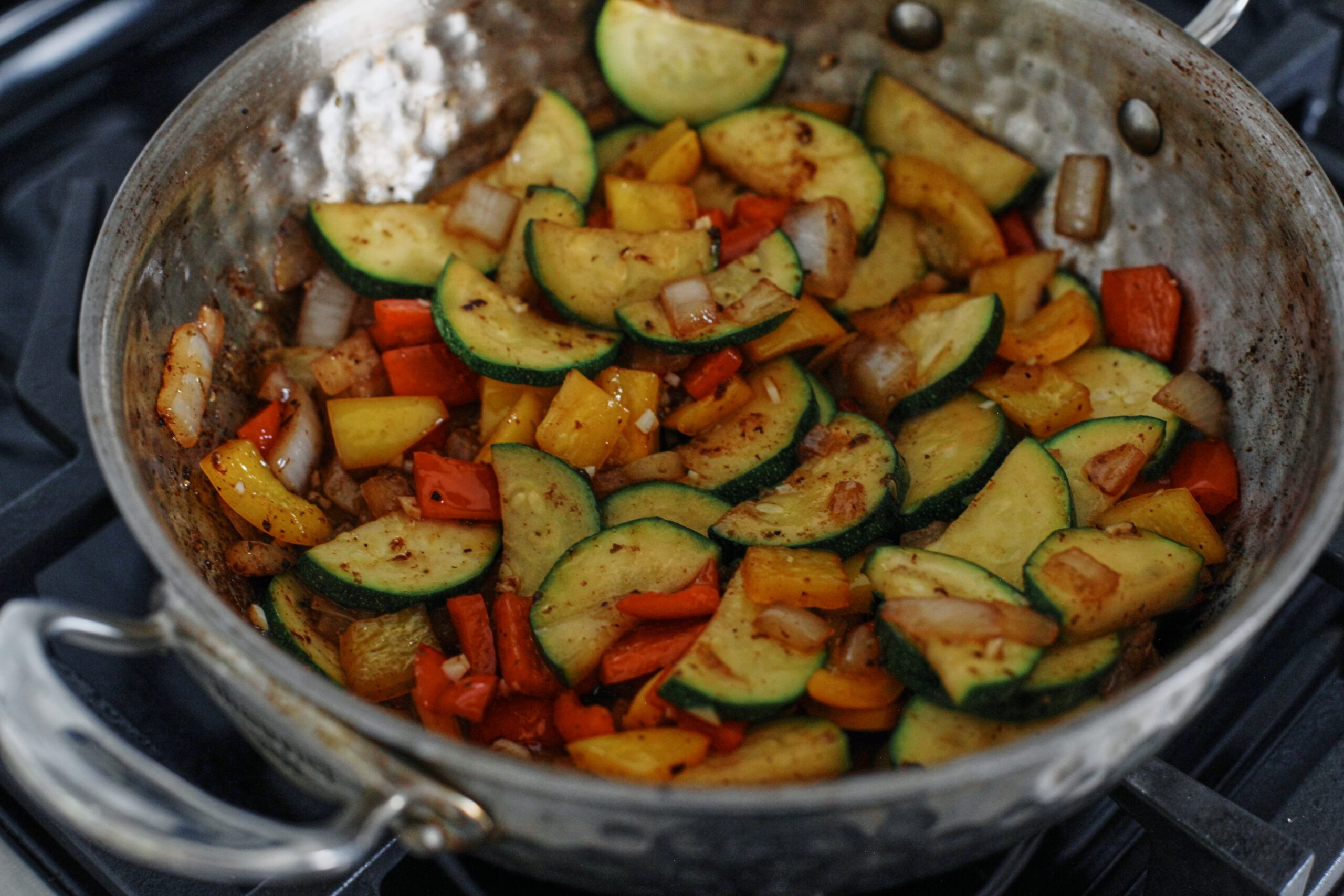 onion, bell peppers, and zucchini being sautéed in a pan