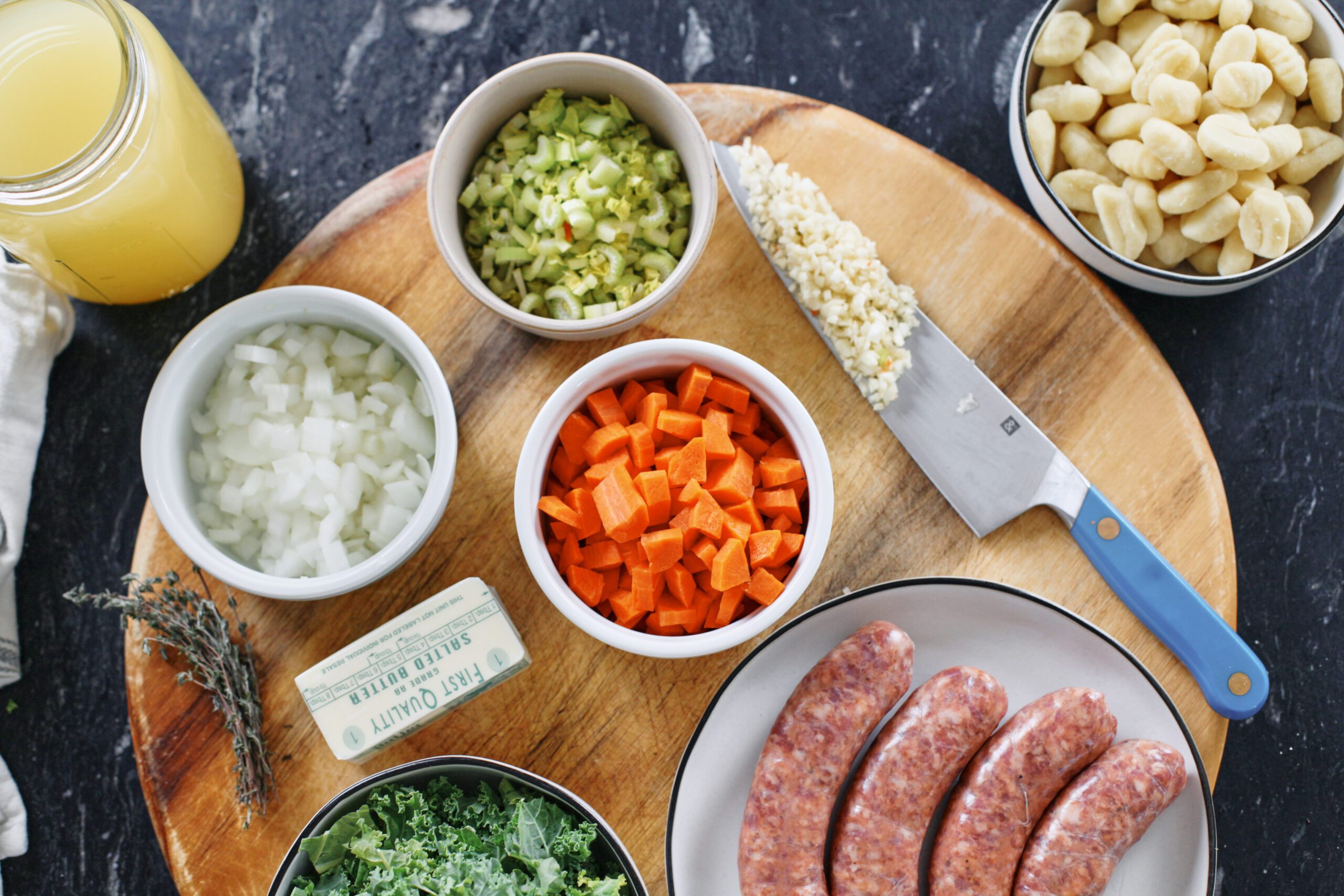 prepped ingredients on a wood cutting board: sausages, chopped carrots, celery, onion, and kale, 1/2 stick of butter, thyme, and garlic on a blue-handled chef's knife