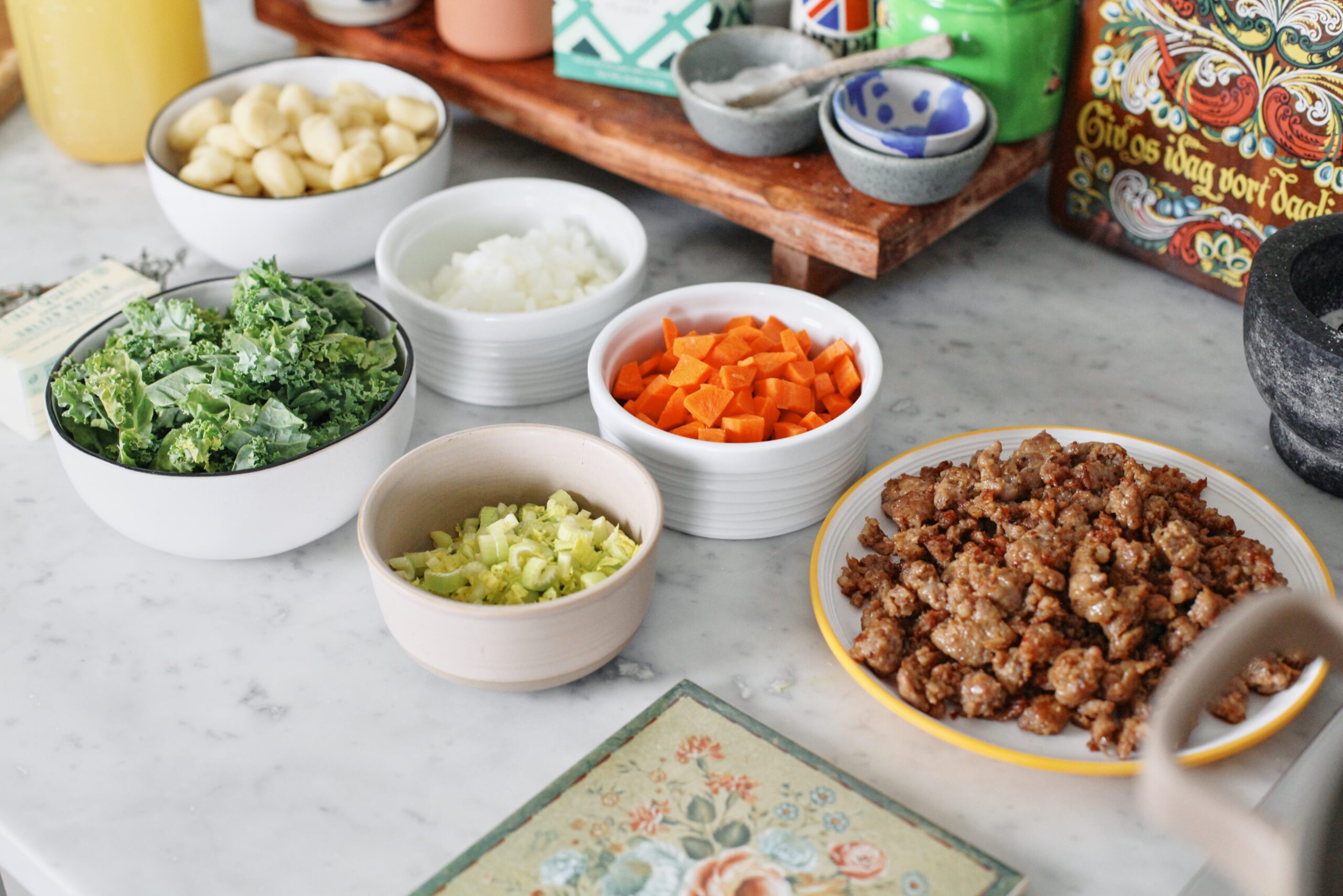 prepped ingredients in bowls: browned sausage, chopped carrots, celery, onion, and kale, and a bowl of gnocchi