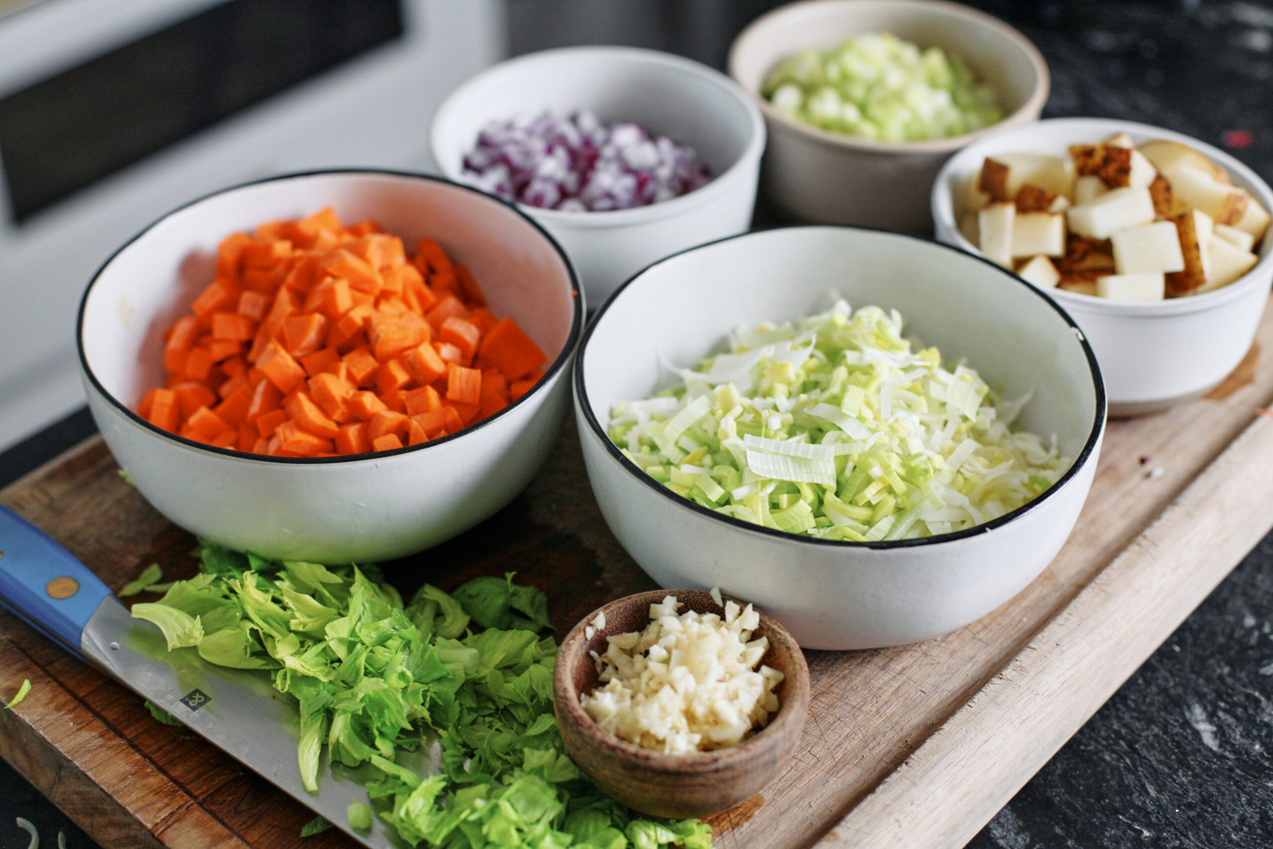 prepped ingredients in bowls on a wood cutting board: chopped carrots, red onion, celery, potato, leeks, garlic, and then celery leaves on the cutting board