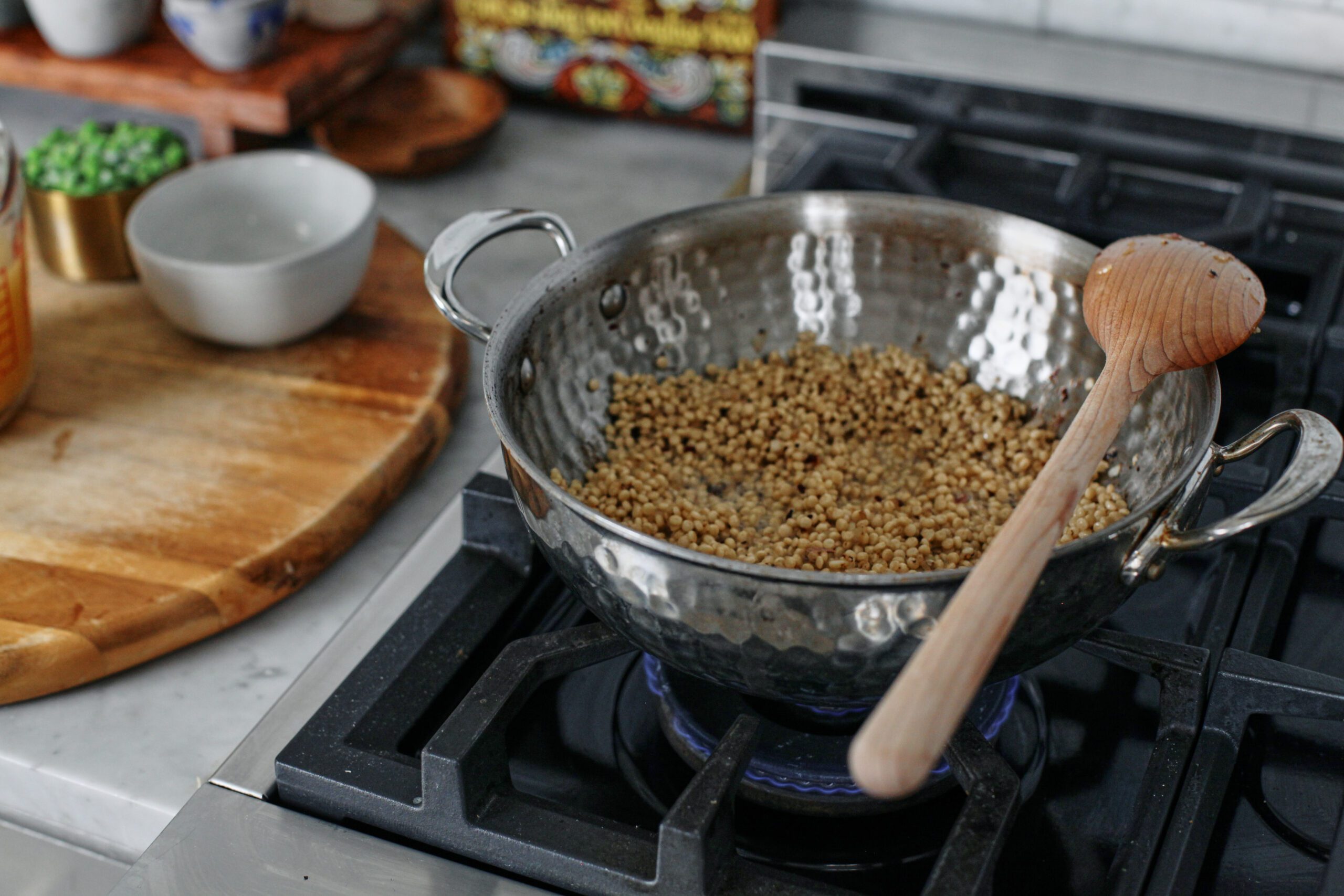 pearl couscous toasting in a sauté pan on the stove, there's a wooden spoon resting across the top of the pan