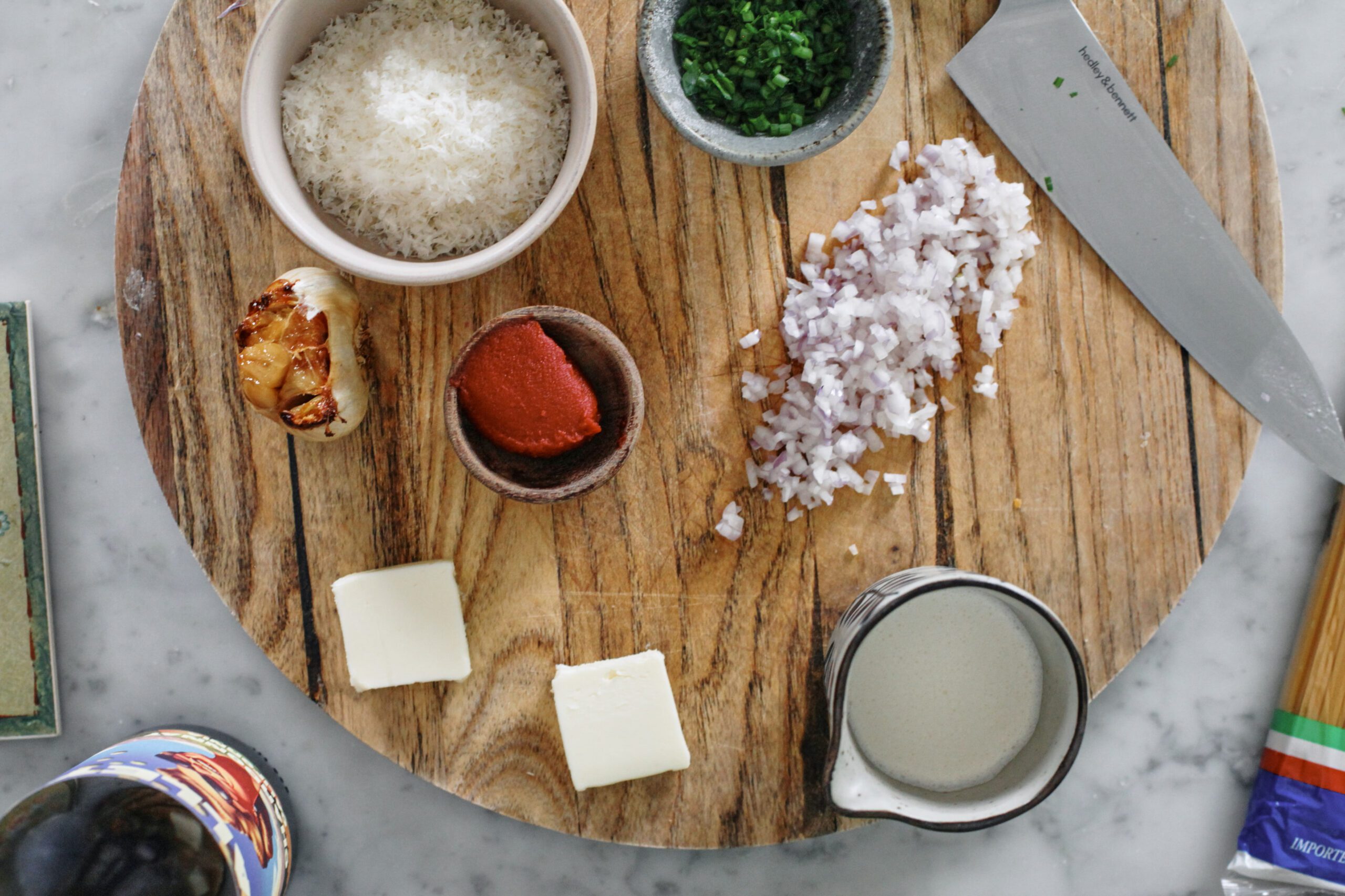 prepped ingredients for roasted garlic spaghetti on a wooden cutting board: grated parmesan cheese in a small bowl, chopped chives in a small bowl, head of roasted garlic, tomato paste in a small bowl, minced shallot, two knobs of butter, a small carafe of heavy cream