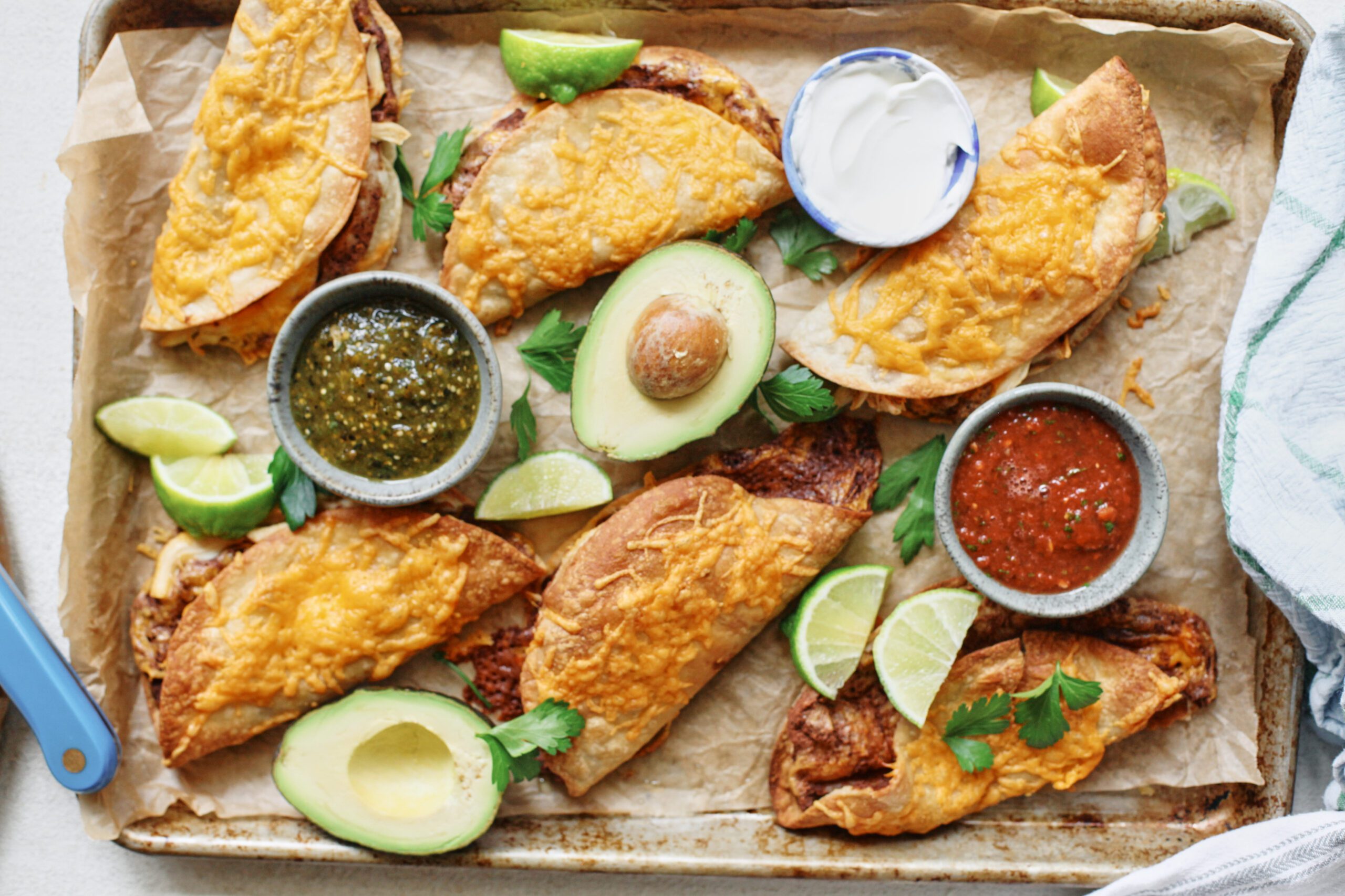 black bean sheet pan tacos arranged on a sheet pan with bowls of red and green salsa, lime wedges, a halved avocado, and cilantro