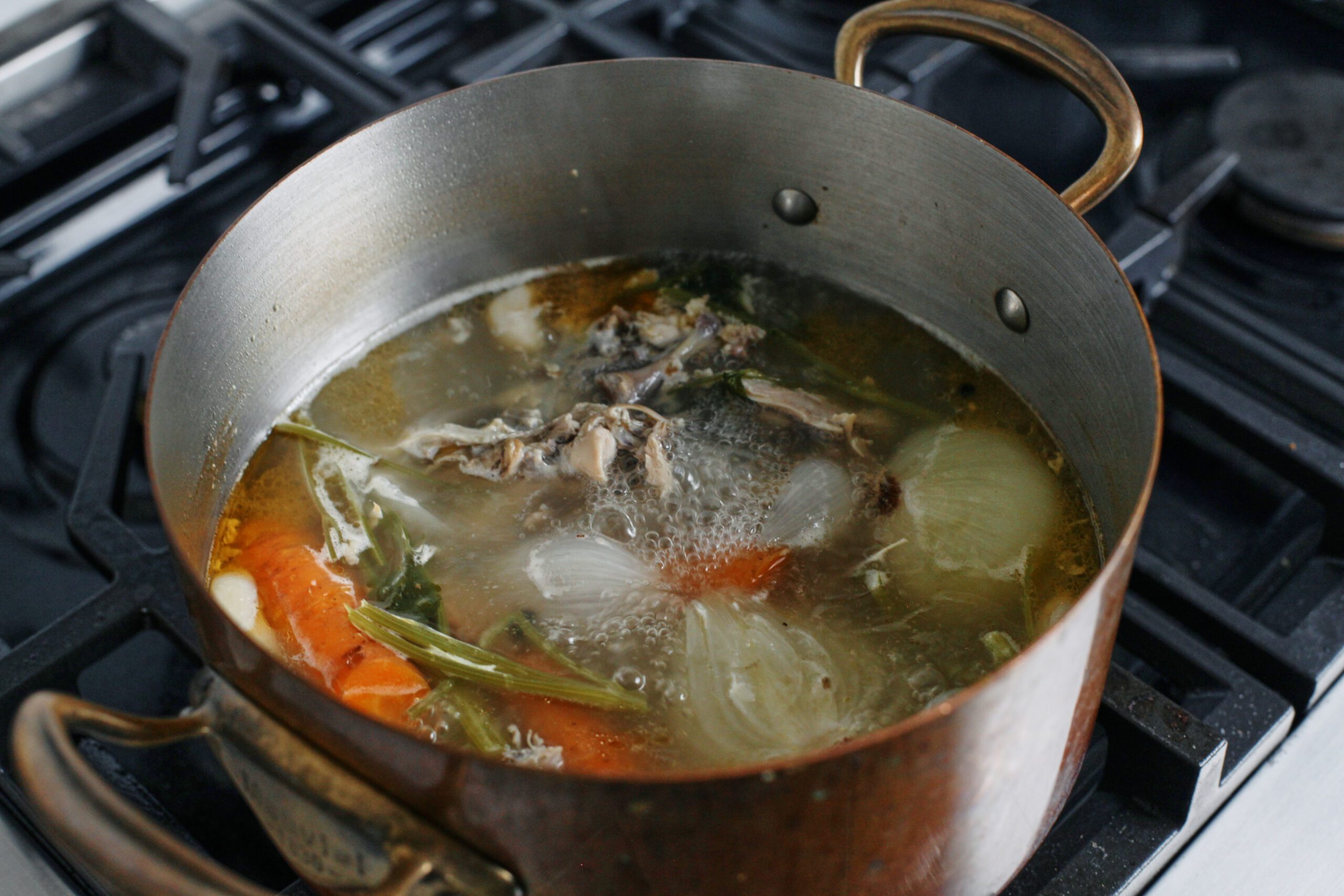 Overhead photo of a large pot of stock simmering on the stove with a rotisserie chicken carcass, onion halves, carrot, celery, ginger, and herbs visible in the broth as it gently bubbles