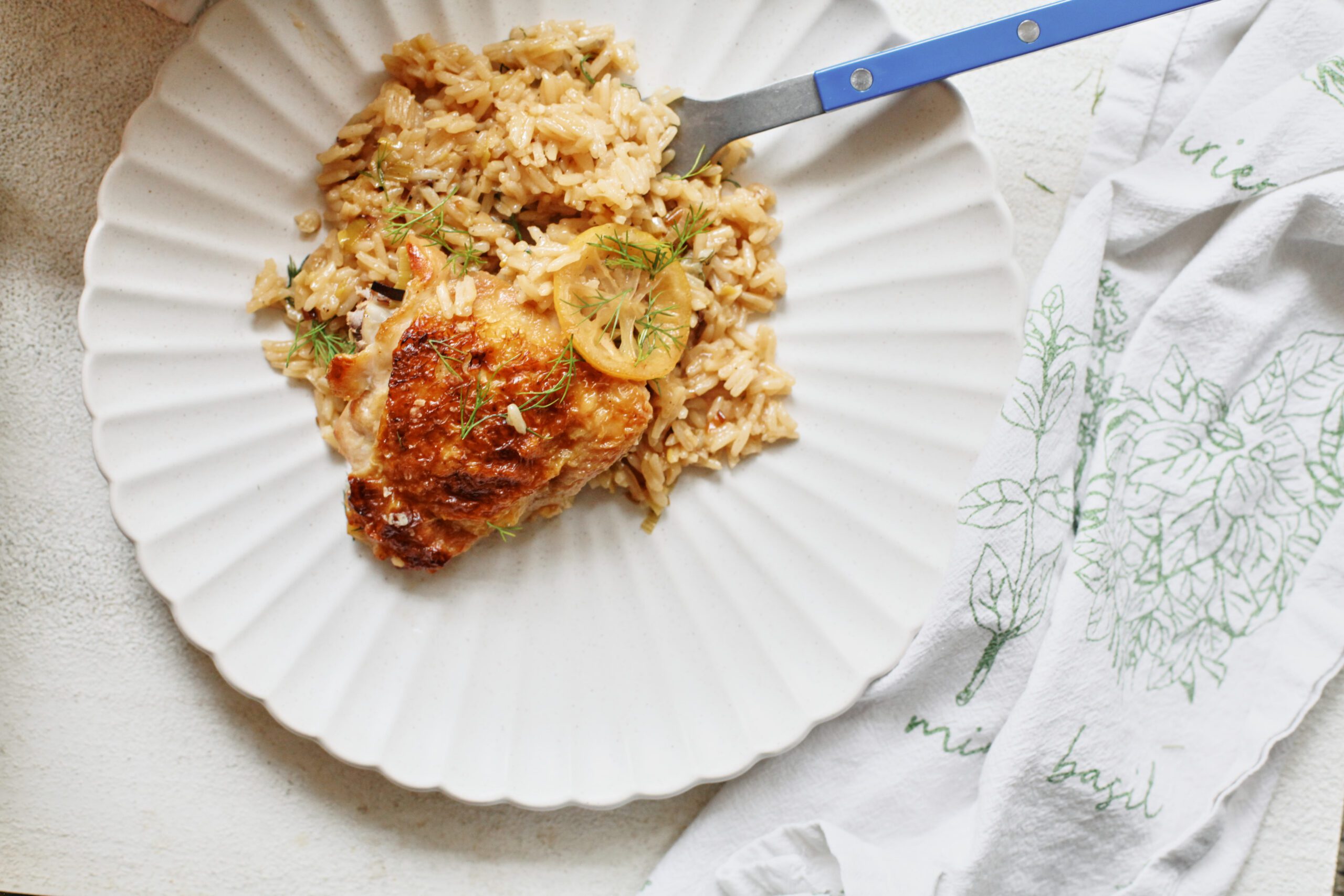 a chicken thigh served over rice, garnished with a seared lemon slice and fresh dill, served on a white plate with scalloped edges