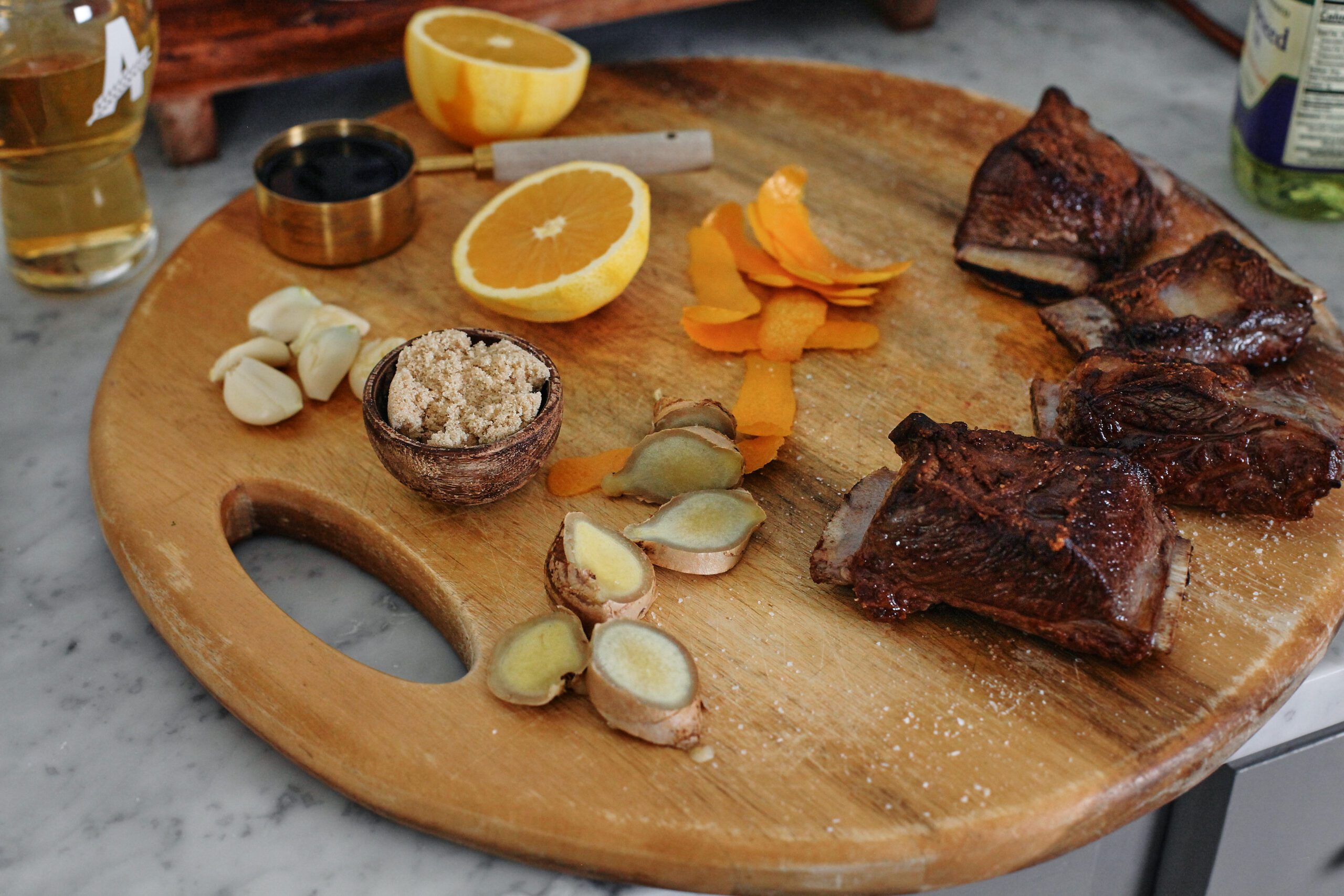 prepped ingredients: garlic cloves, orange peels, an orange, 1/2 cup of soy sauce, ginger sliced into coin-shaped pieces, seared short ribs