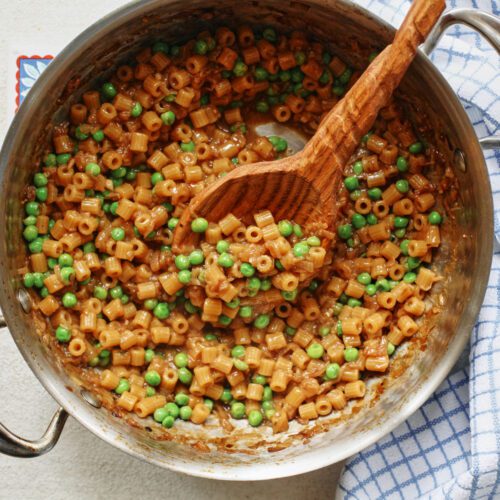 overhead photo of caramelized shallot ditalini with peas in a sauté pan with a wooden spoon
