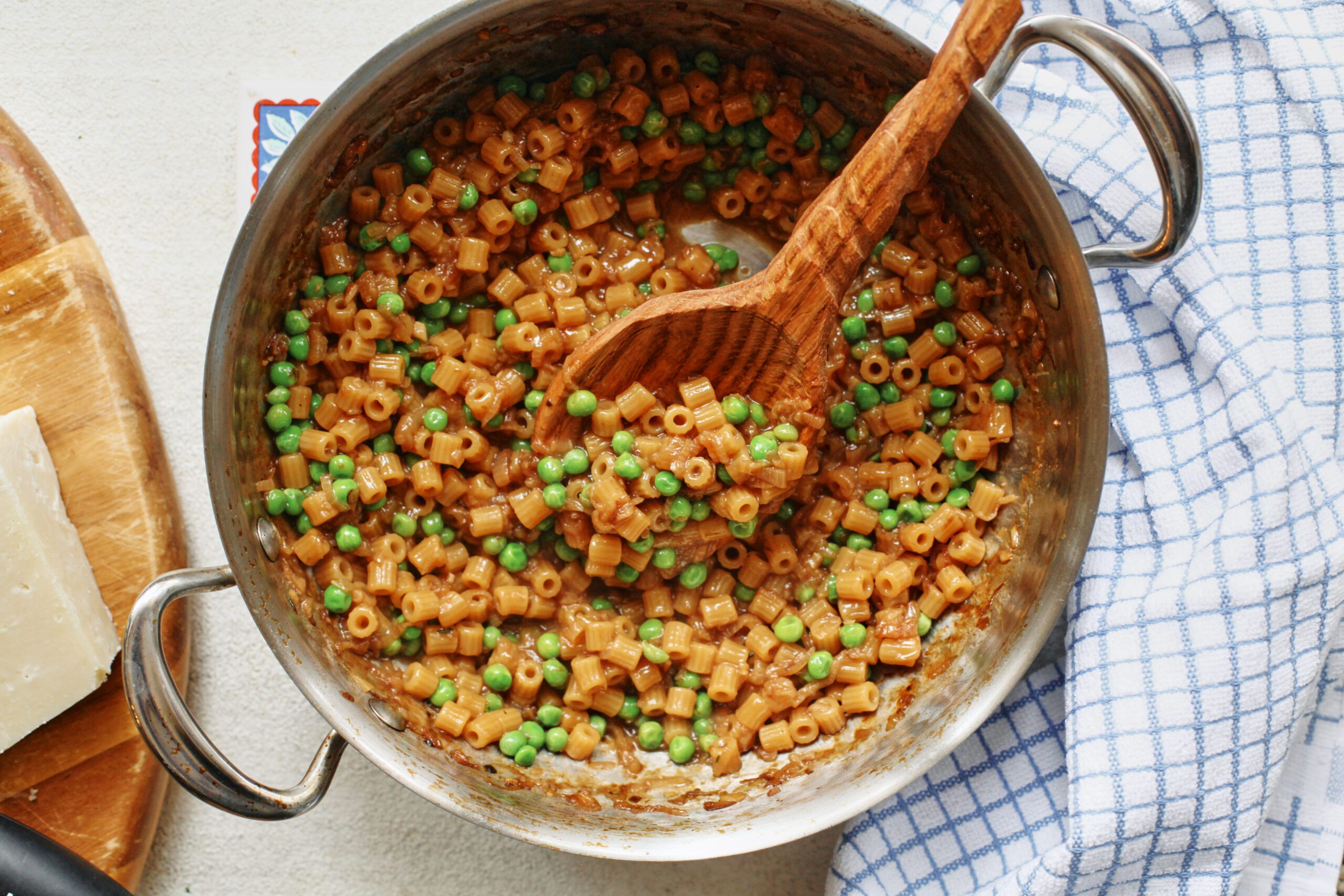 overhead photo of caramelized shallot ditalini with peas in a sauté pan with a wooden spoon