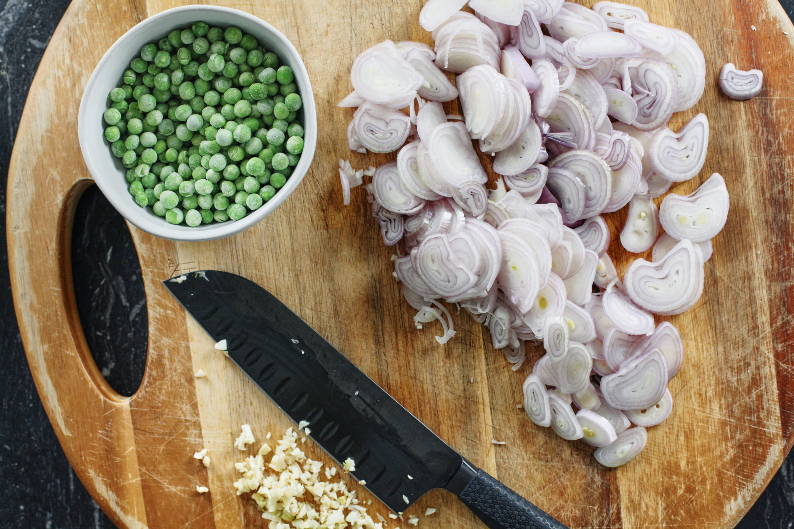 overhead photo of a bowl of frozen peas, sliced shallots, minced garlic, and a knife on a round wooden cutting board